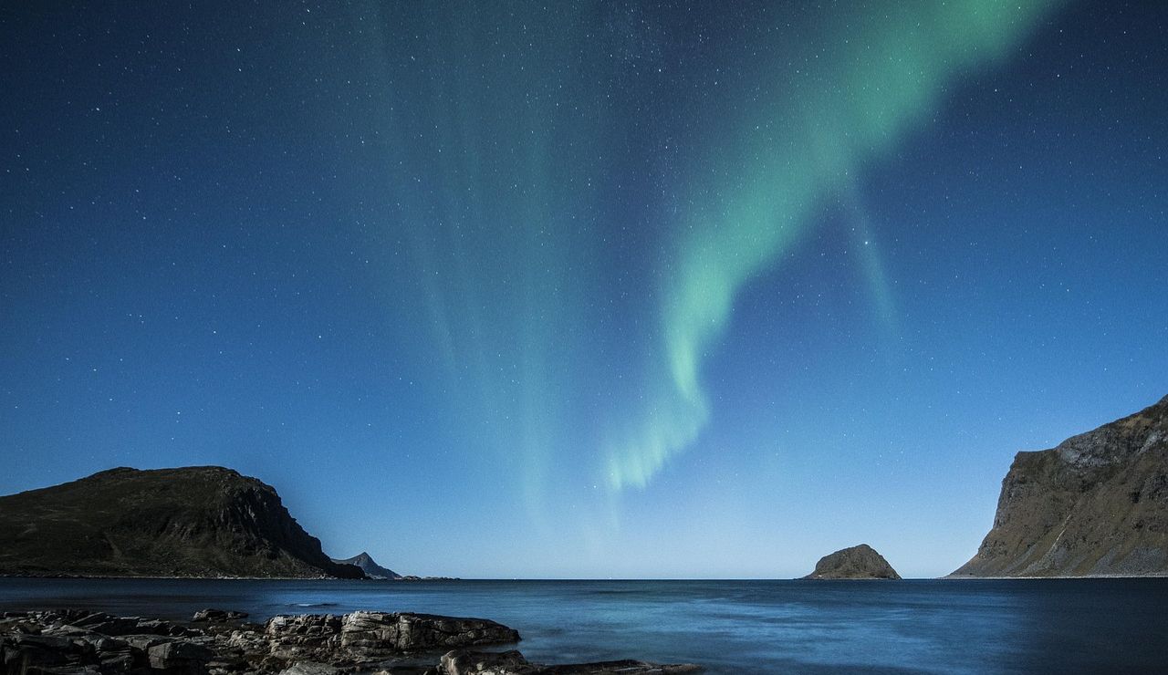 Northern lights streaking across a starry sky above rocky coastline and calm waters in Senja, Norway.