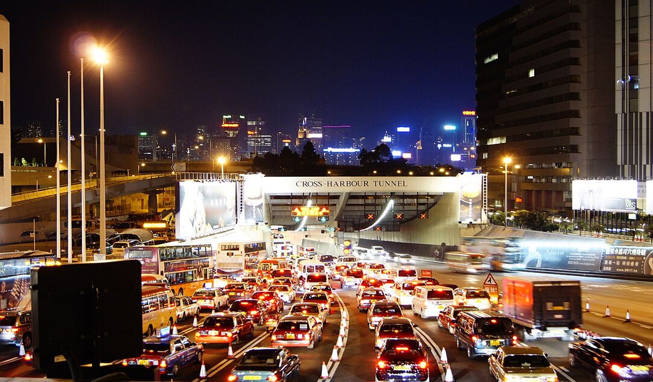The image shows a dense night-time traffic jam at the entrance of an illuminated urban tunnel, surrounded by high-rise buildings and bright city lights.