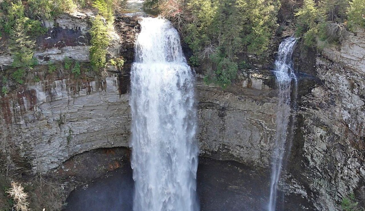 A wide, powerful waterfall drops from a rocky cliff beside a narrow stream, all framed by lush forest.