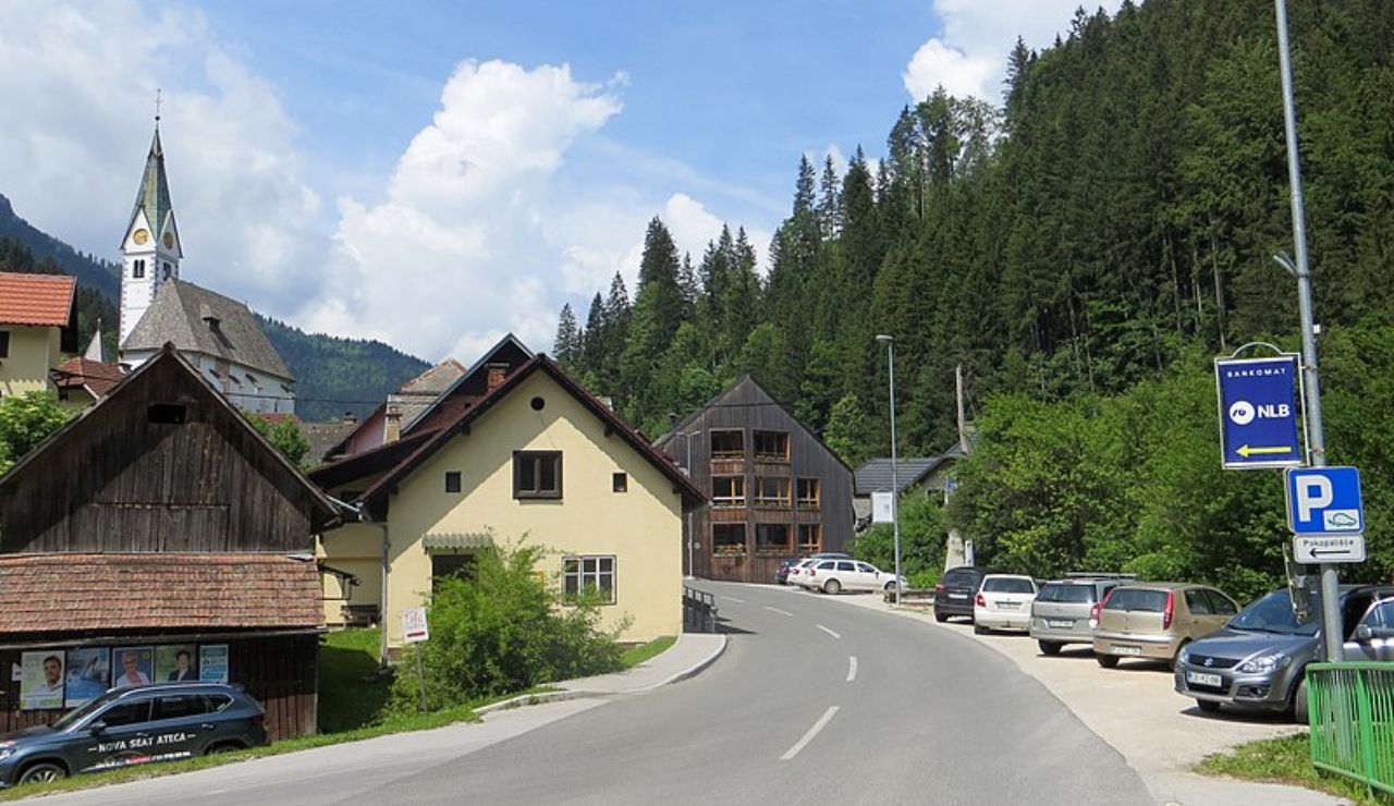 cenic mountain village of Solčava, Slovenia, with alpine houses, a church steeple, forested hills, and parked cars along a winding road.