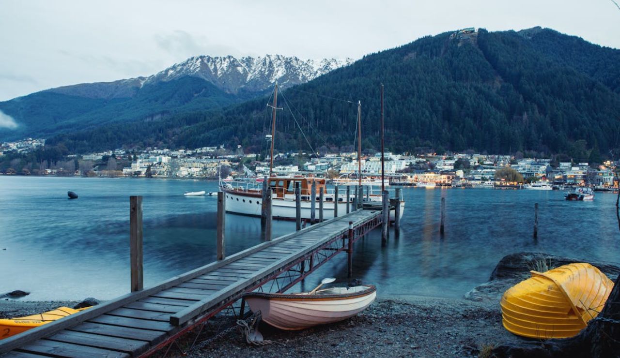 Scenic view of Lake Wakatipu and Queenstown, New Zealand, with boats, a wooden dock, and snow-capped mountains in the background.