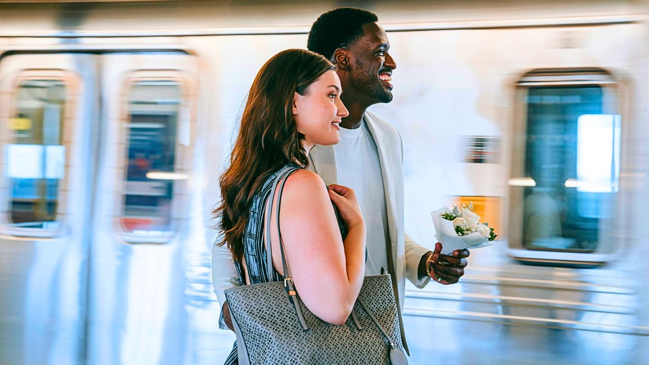 A couple with small suitcases smiling on a Spanish train platform, ready to board a high-speed train.