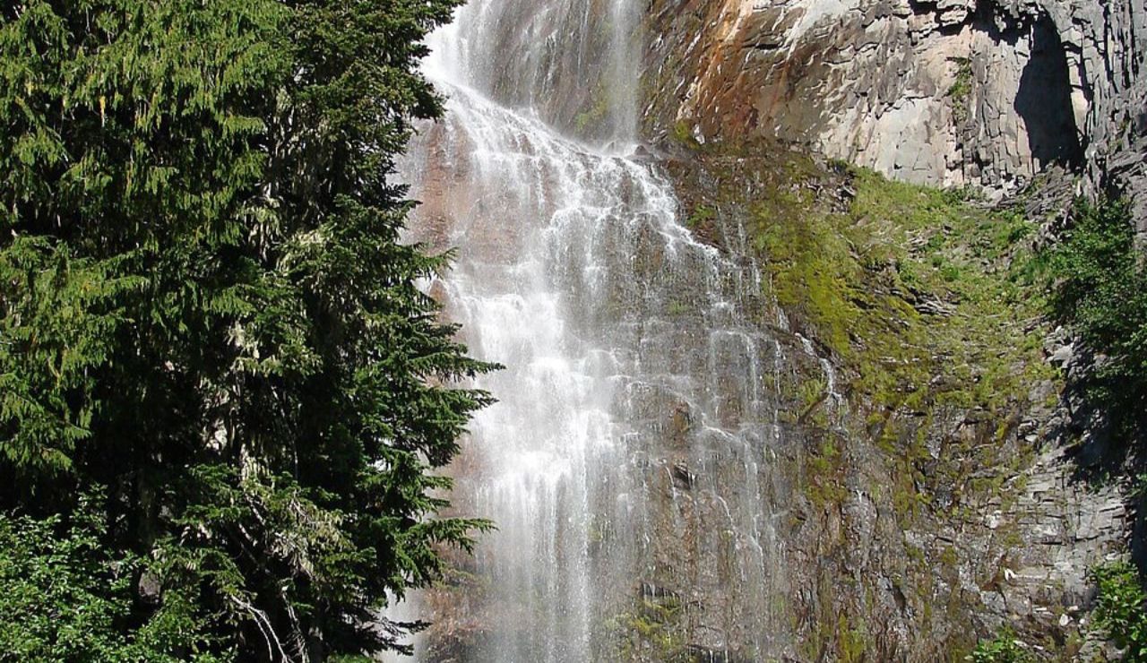 A gentle cascade flows down a rugged, mossy cliffside, framed by tall evergreens and jagged rock formations.