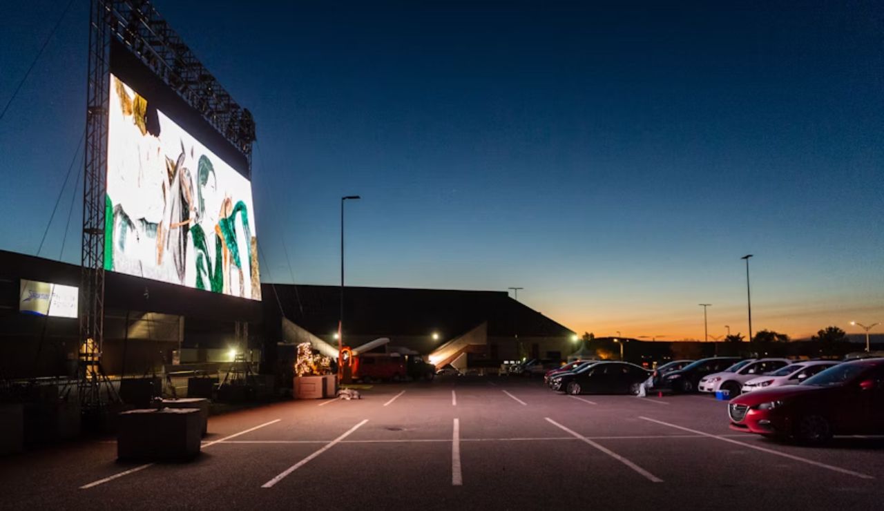 Drive-in movie theatre at dusk with cars parked facing a large outdoor screen.
