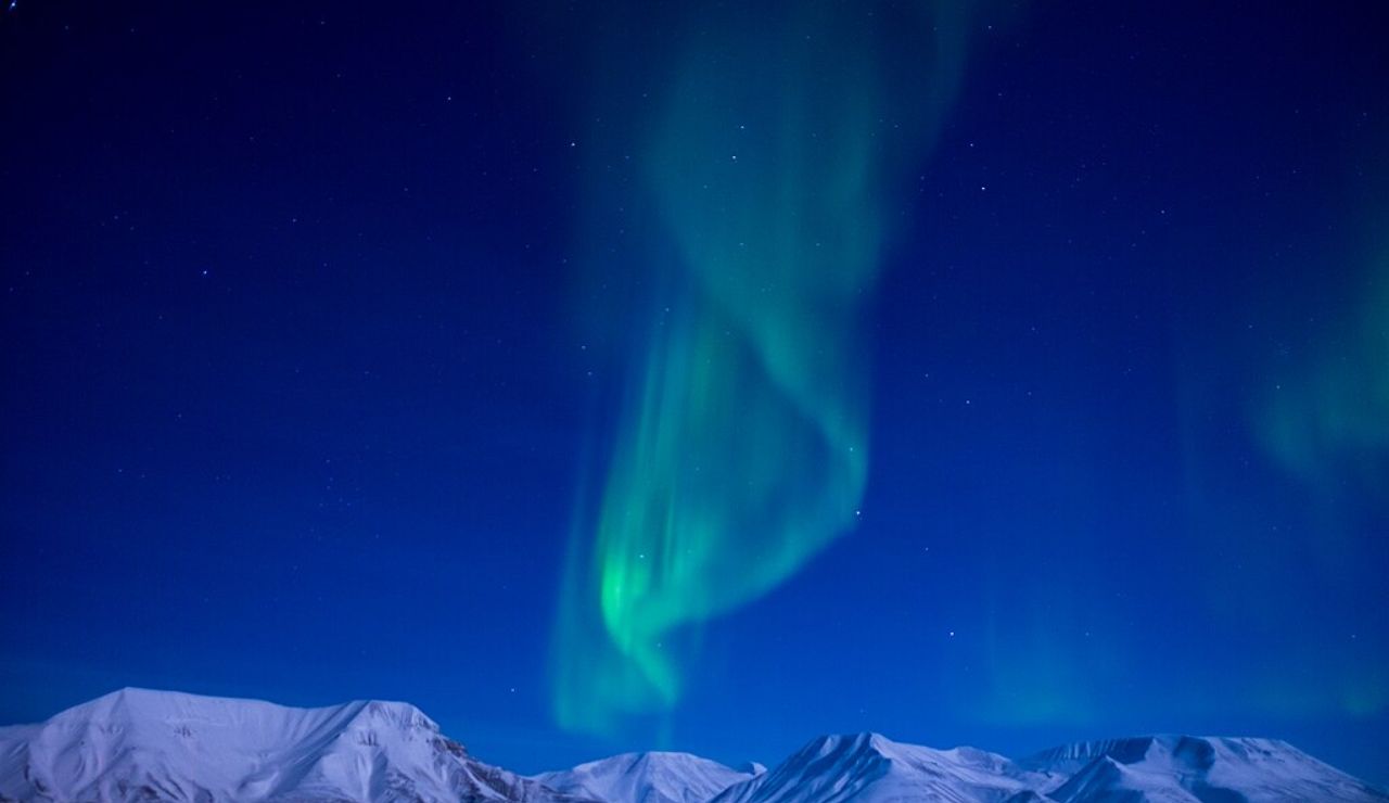 Northern lights swirling above snow-covered mountains under a starry sky in Svalbard, Norway.