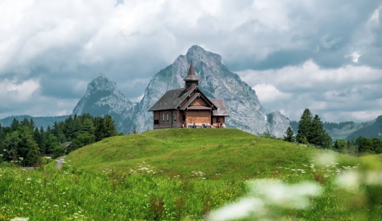 Charming wooden chapel on a grassy hill in the Swiss Alps, Switzerland, with dramatic mountains and cloudy skies in the background.