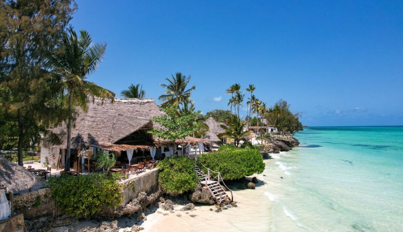Thatched beachside resort on the turquoise shores of Zanzibar, Tanzania, with palm trees and clear blue skies.
