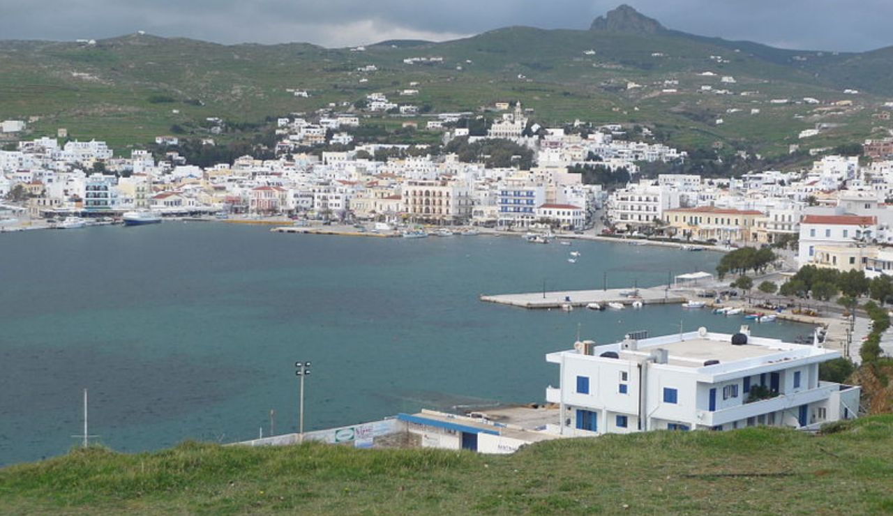 View of Tinos Town and harbor on Tinos Island, part of the Cyclades in Greece, with white buildings and green hills in the background.