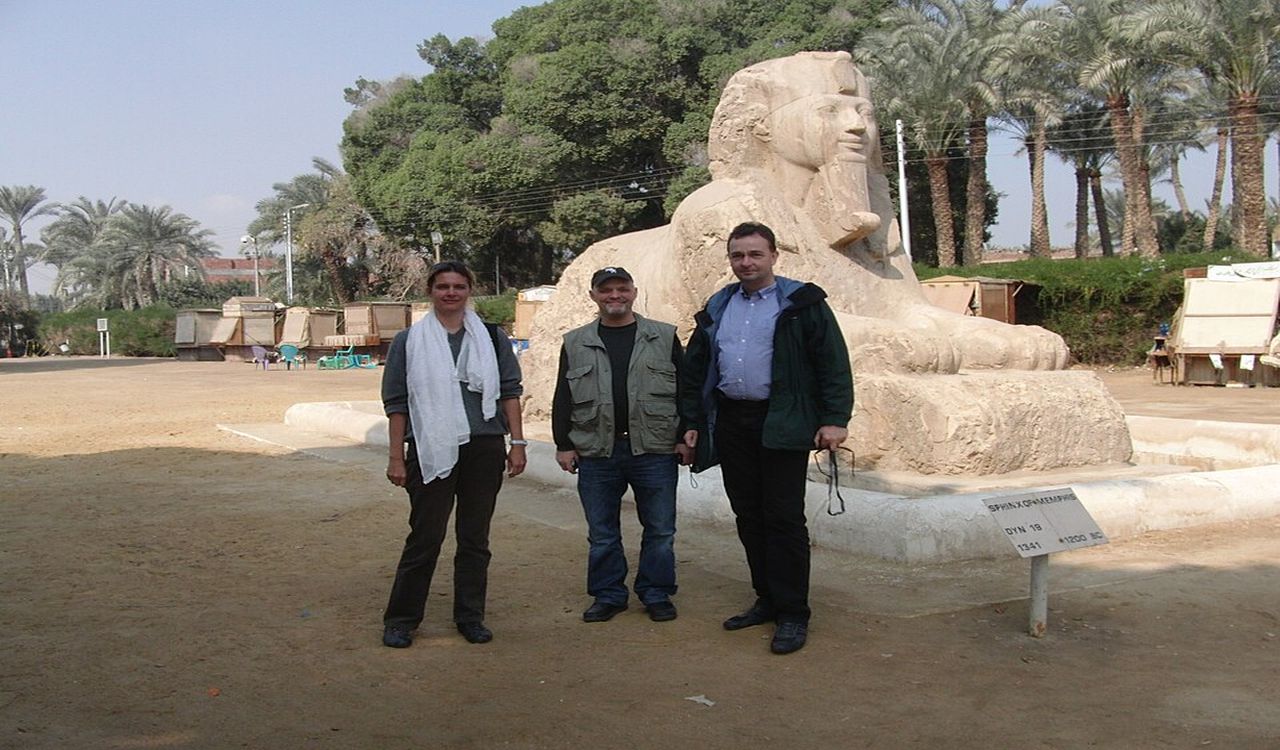 Three people pose in front of an ancient sphinx statue, surrounded by palm trees and a sandy open area with market stalls nearby.