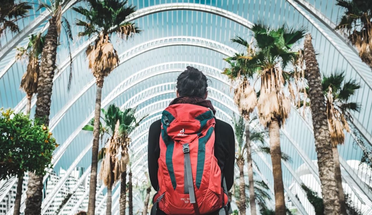 Traveler with red backpack standing under modern architectural arches and palm trees.