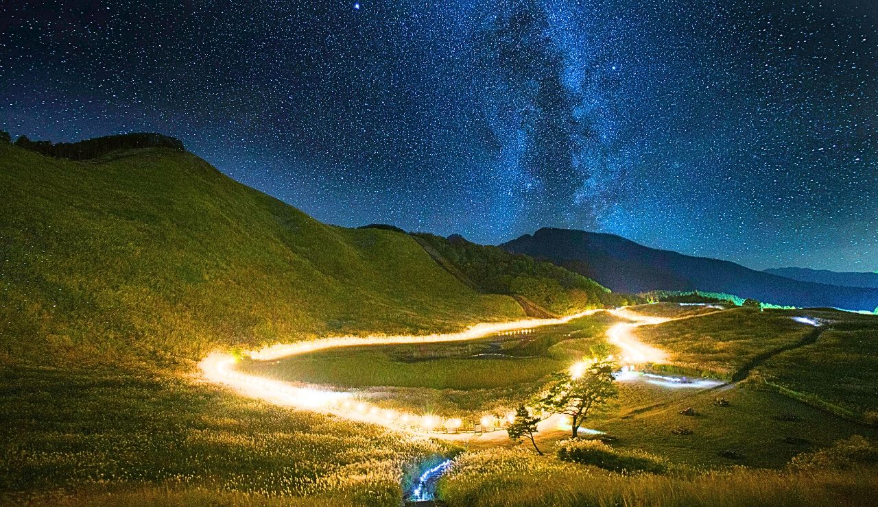 A winding, yellow-lit road curves through grassy hills beneath a clear, starry sky and the Milky Way.