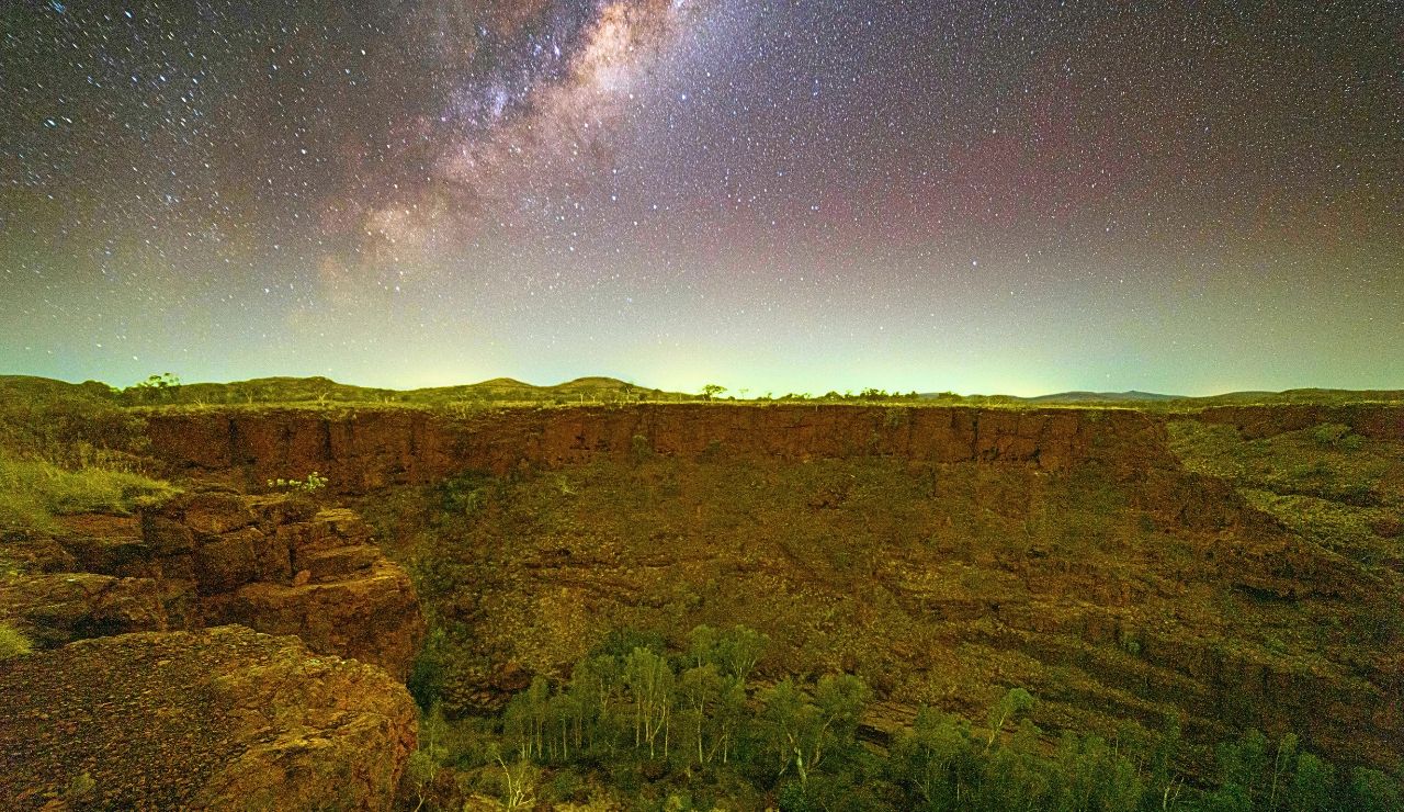 A deep canyon with rocky cliffs and sparse vegetation lies beneath a star-filled night sky and the Milky Way.