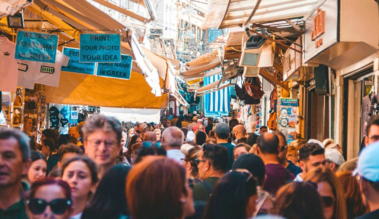 A crowded outdoor market features people walking among stalls beneath English and Greek signs, with goods displayed under awnings.