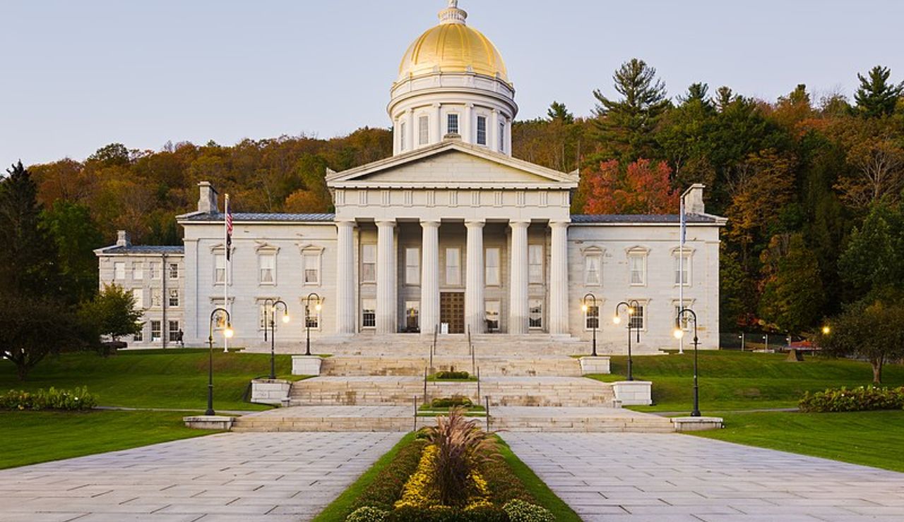 Front view of the Vermont State House in Montpelier with its golden dome, neoclassical columns, and surrounding autumn foliage during early evening.