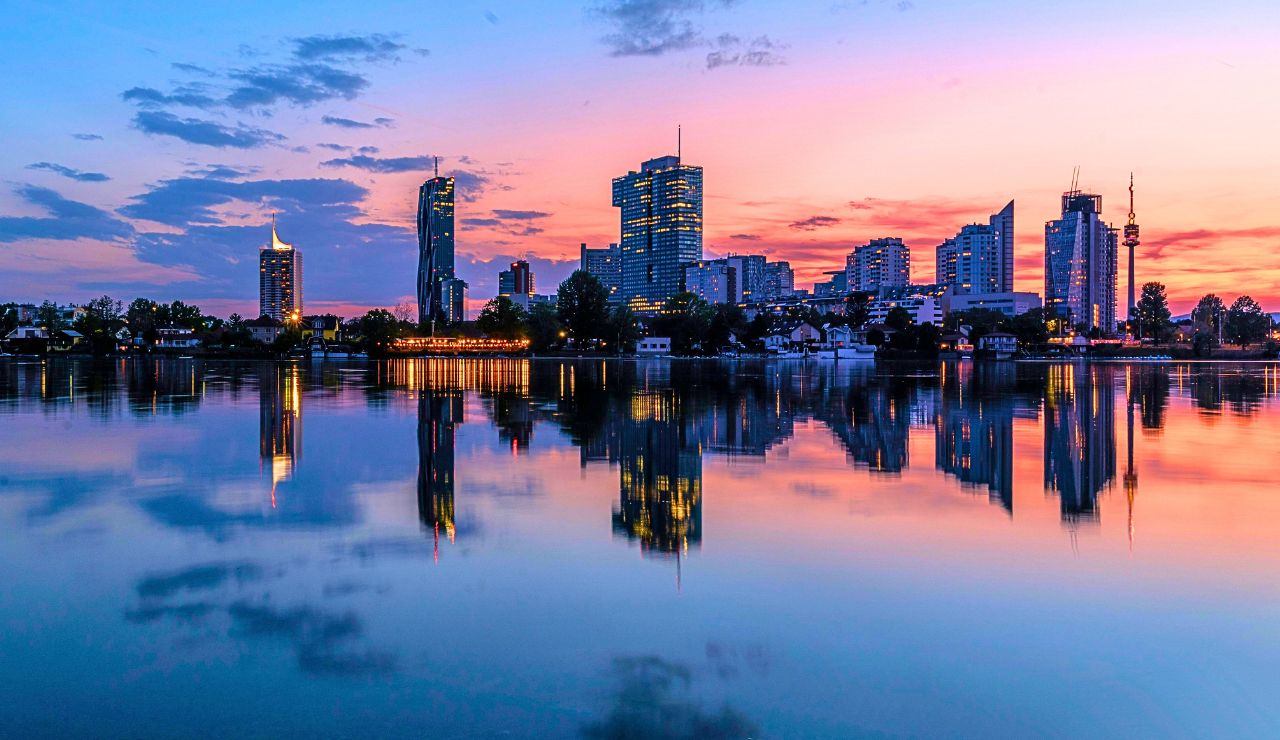 Modern high-rise buildings of the city skyline are reflected in a calm river at sunset beneath pink, orange, and blue skies.
