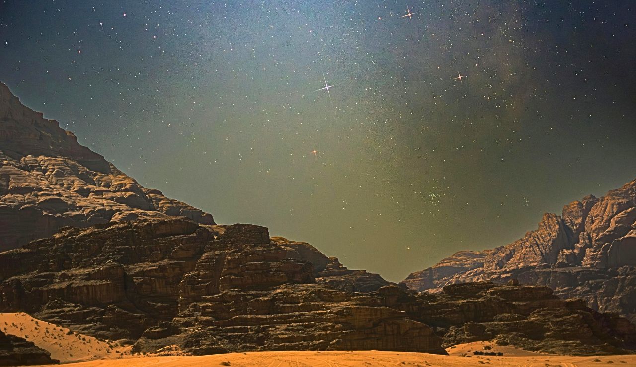 A rocky desert with sand dunes and rugged cliffs sits beneath a star-filled night sky.