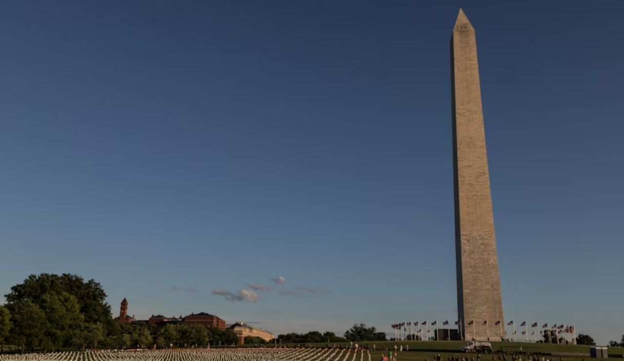 Washington Monument towering over the National Mall in Washington, D.C., with white flags arranged on the grass and a clear blue sky overhead