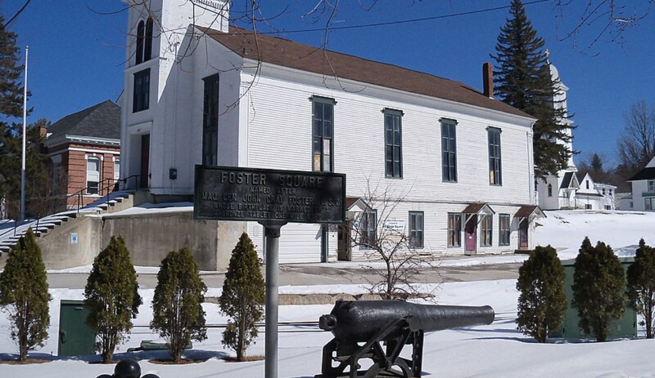 Historic white church building in snowy Foster Square with Civil War cannon and commemorative plaque in Lancaster, New Hampshire.