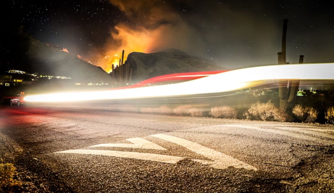 Nighttime wildfire on mountain with streaking car lights and desert cacti.