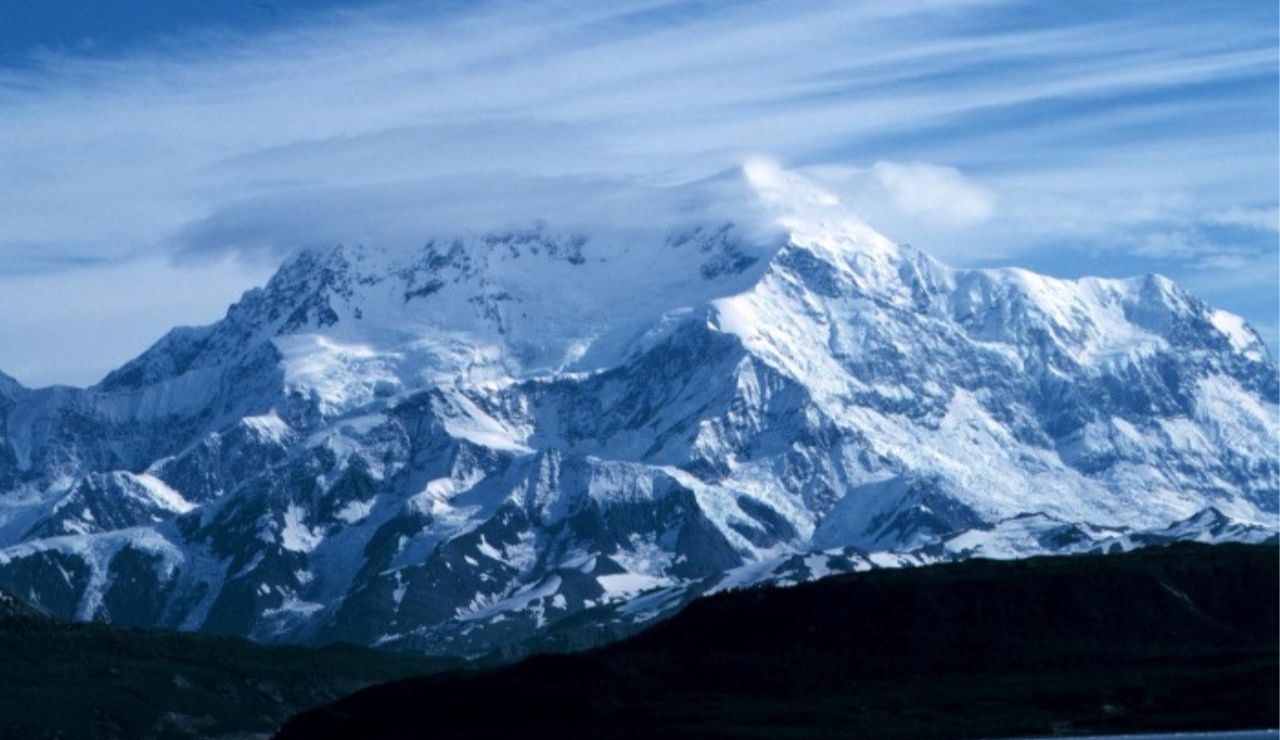 A towering snow-covered mountain rises under streaked clouds, its icy peaks glowing against the deep blue sky.