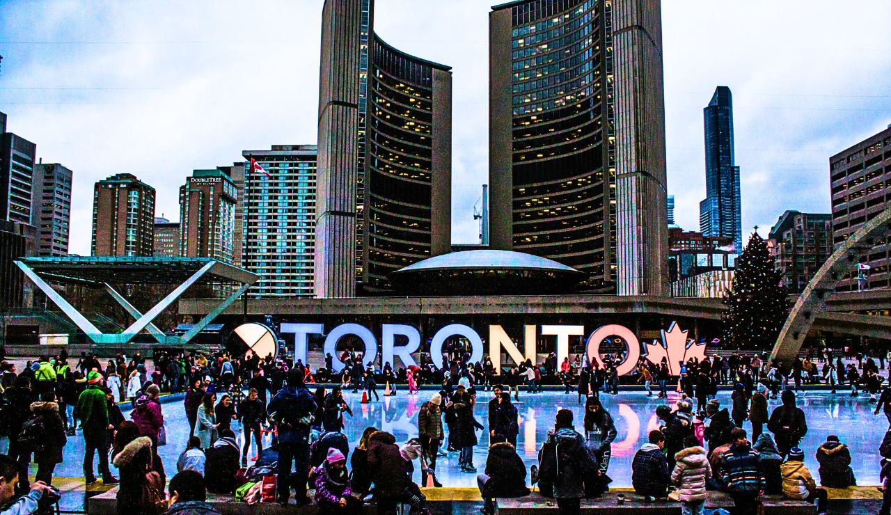 Crowds gather at an outdoor ice rink in Nathan Phillips Square before the Toronto sign and city hall on a cloudy day.