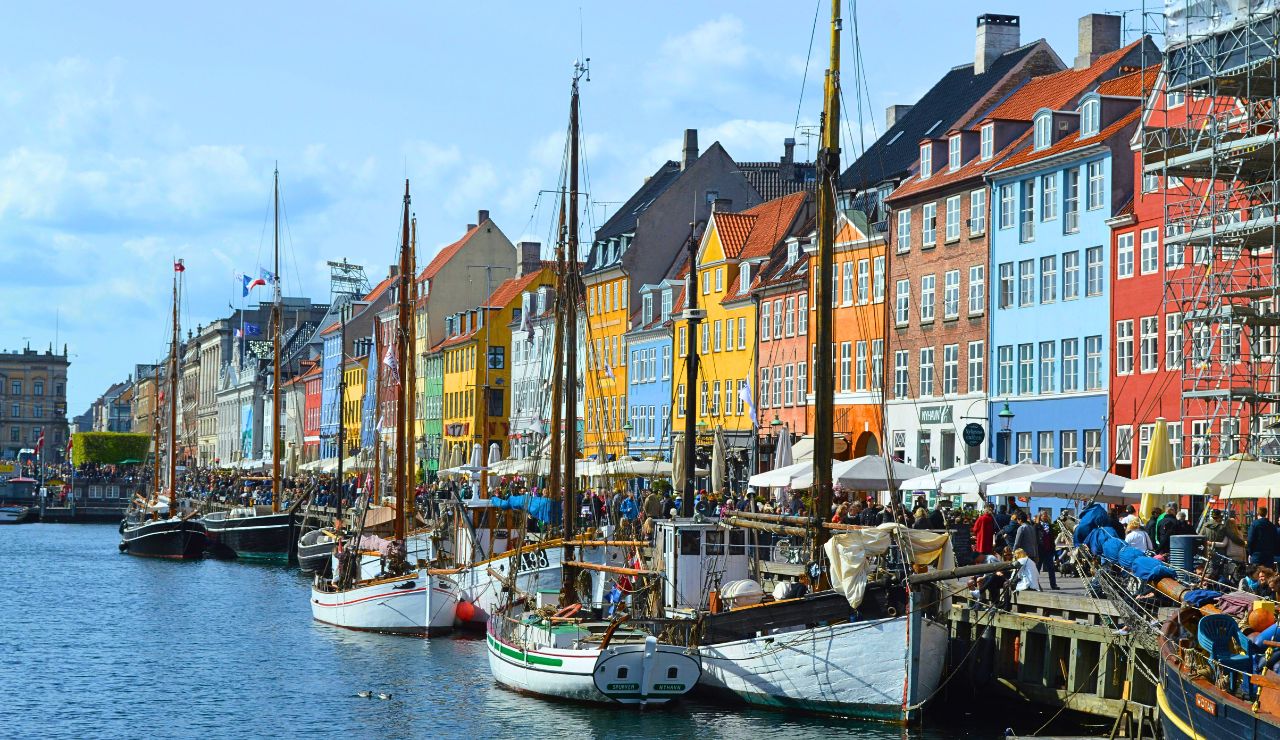 Colorful buildings line a lively canal with docked boats, outdoor cafes, and people enjoying the waterfront.