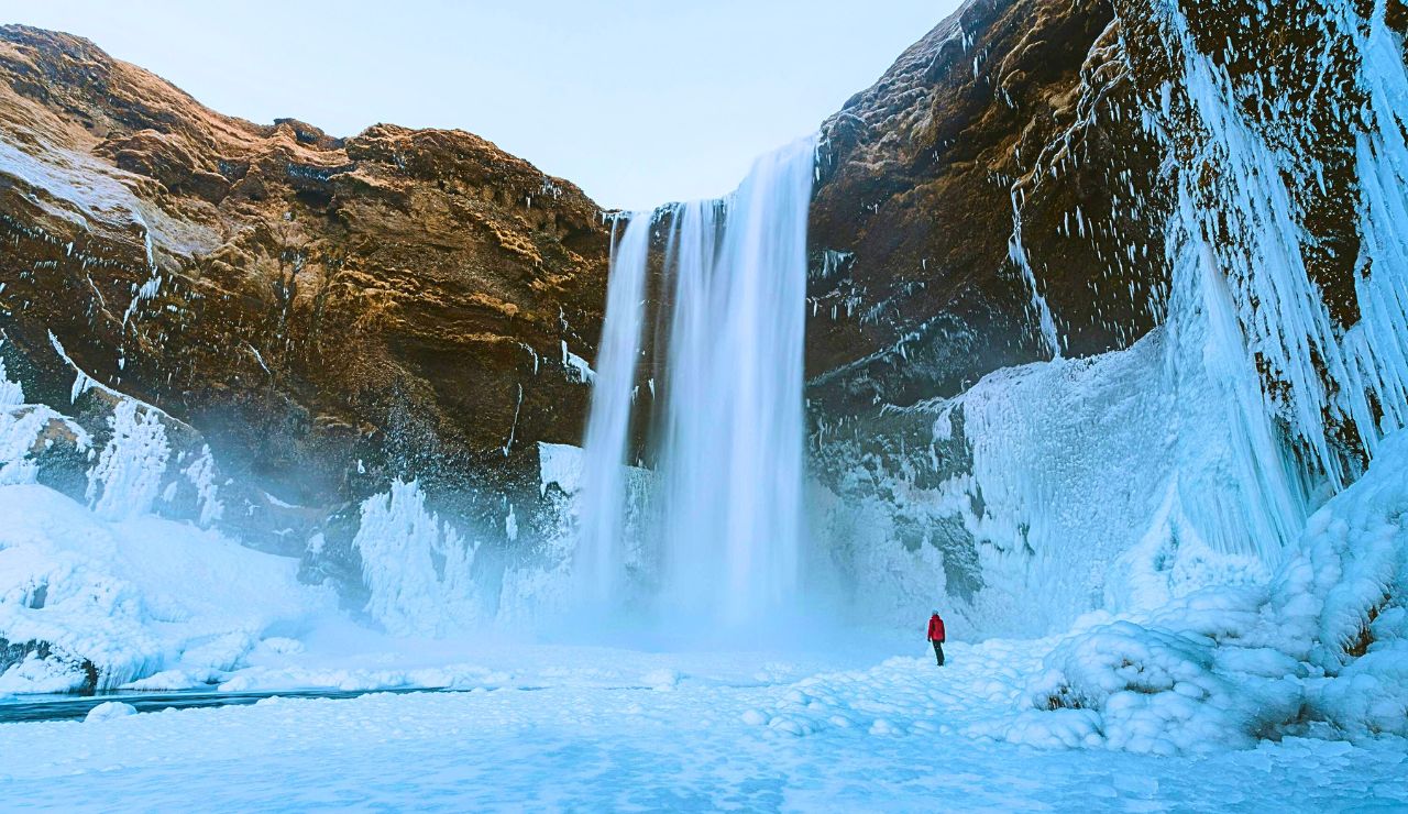 A person in a red jacket stands on ice near a large, snow-covered waterfall and frozen cliffs.