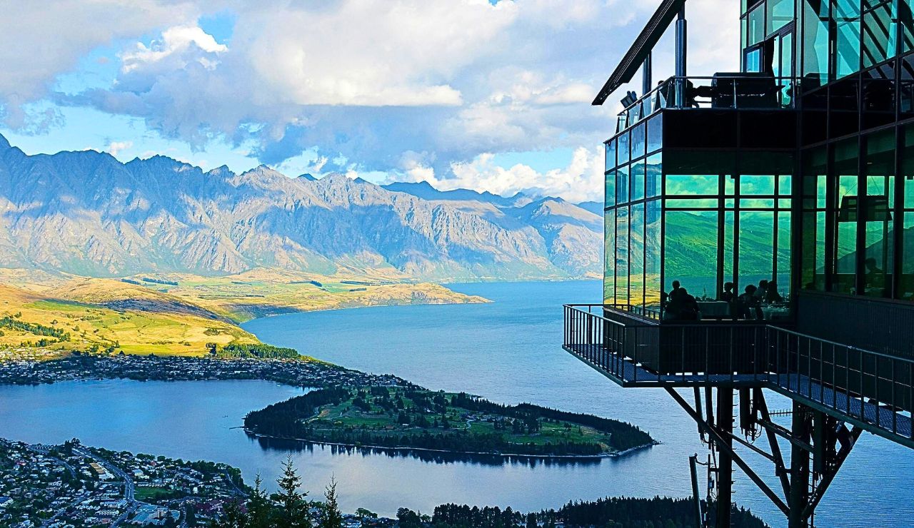 A glass-walled building overlooking a lake, small island, and distant mountains houses several visible occupants beneath a partly cloudy sky.