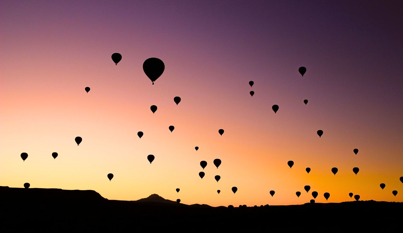 Silhouettes of hot air balloons drift across the sky at dawn or dusk, with mountains on the horizon.