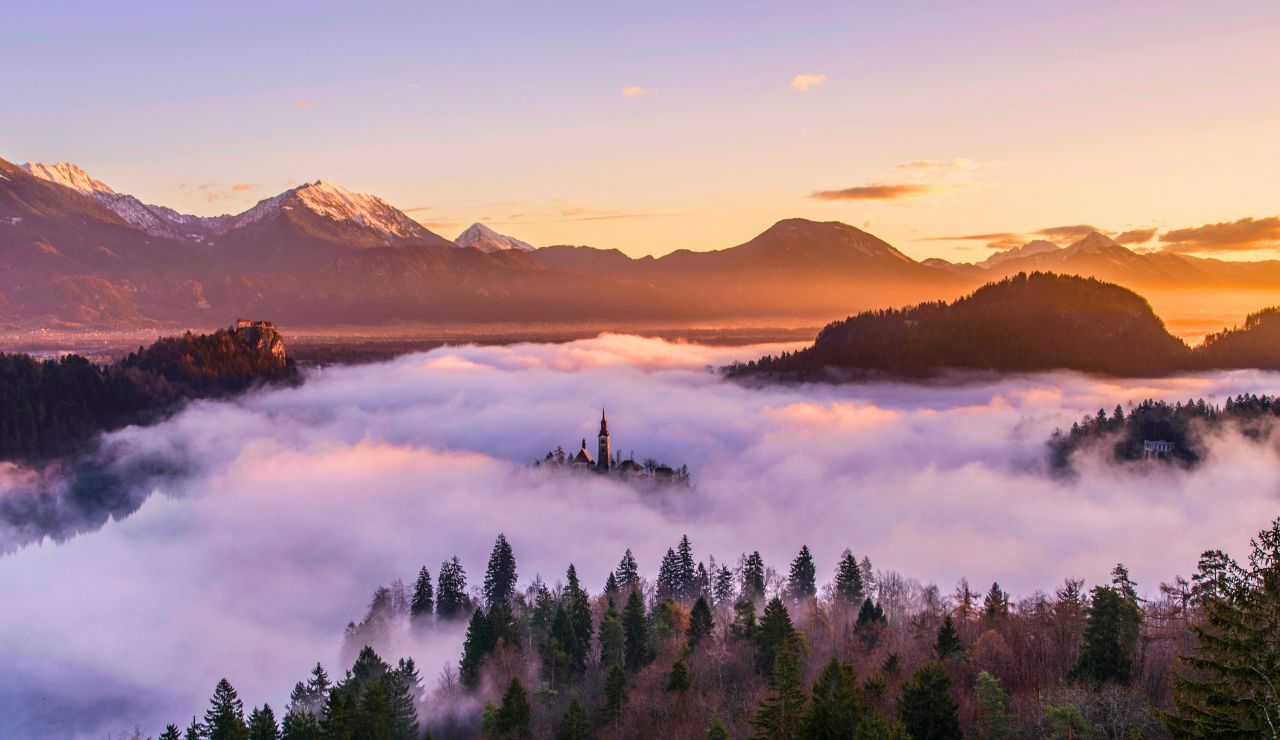 A church stands on an island amid mist and trees, backed by mountains beneath a vibrant sunrise sky.