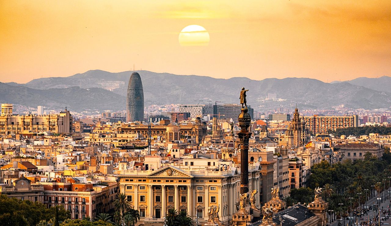 Aerial view of Barcelona at sunset shows historic buildings, the Columbus Monument, and distant mountains beneath a golden sky.