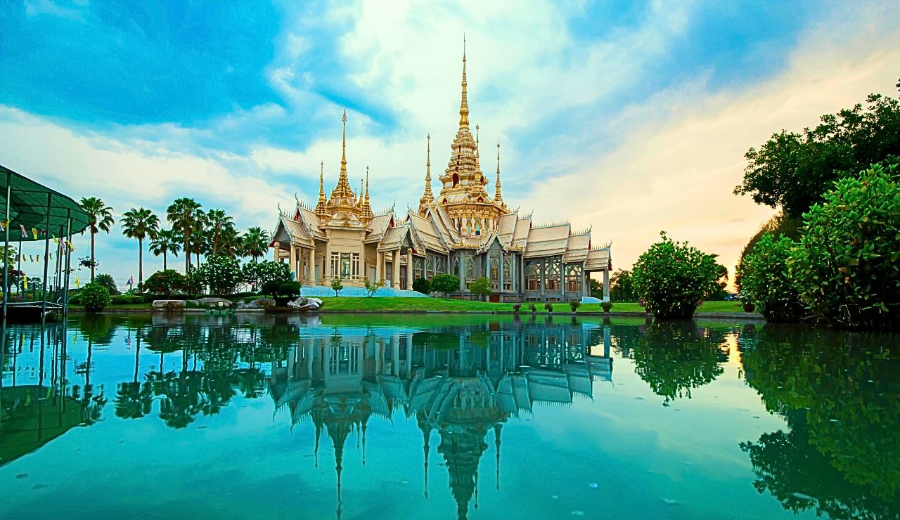 A large, ornate temple with multiple spires stands by a calm pond that reflects the structure and surrounding greenery under a blue sky.