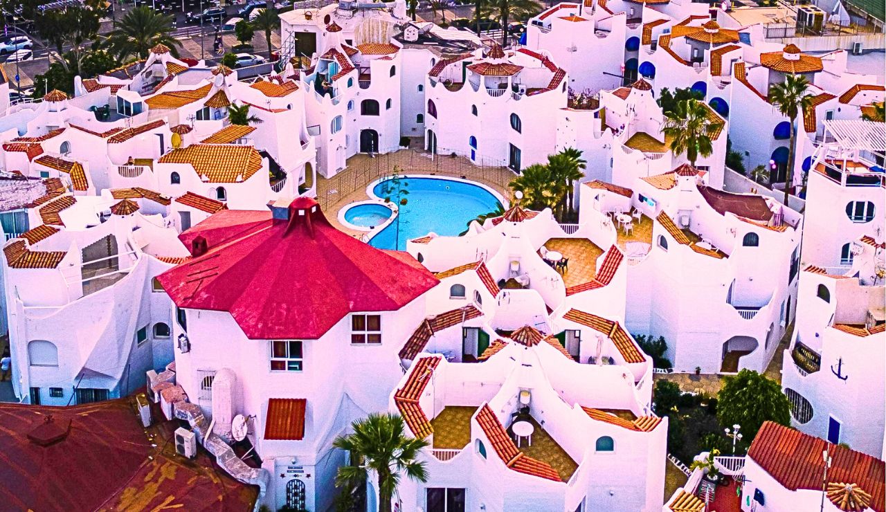 Aerial view of white Mediterranean-style buildings with red roofs encircling a central pool and palm-lined courtyard.