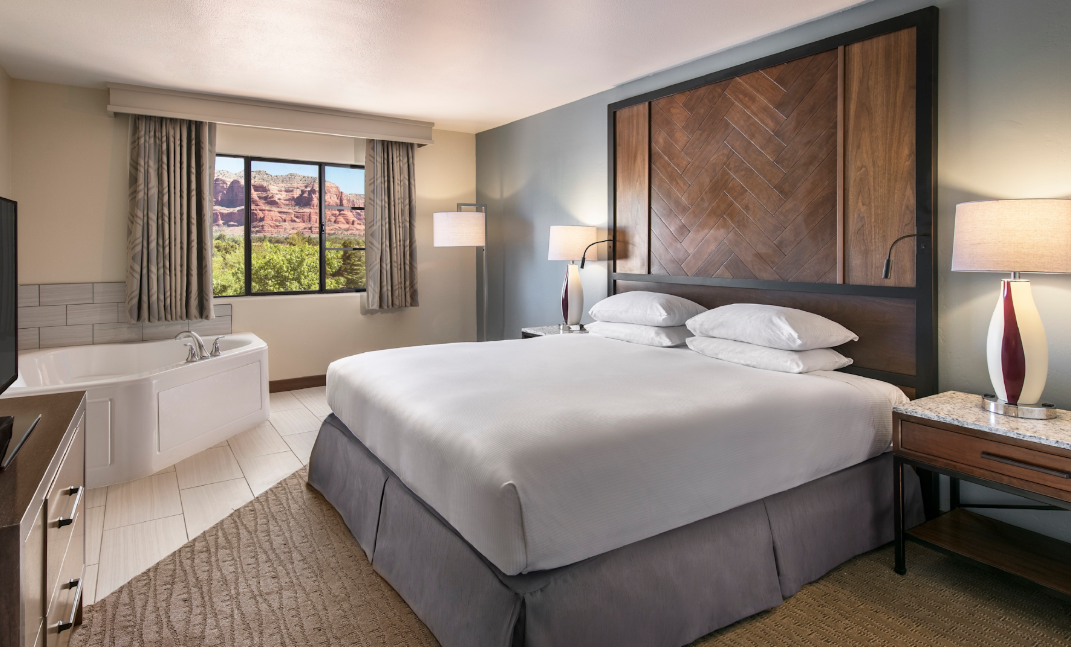 Hotel room with a king bed, two bedside tables with lamps, a large scenic window, and a white bathtub in the corner by the window.