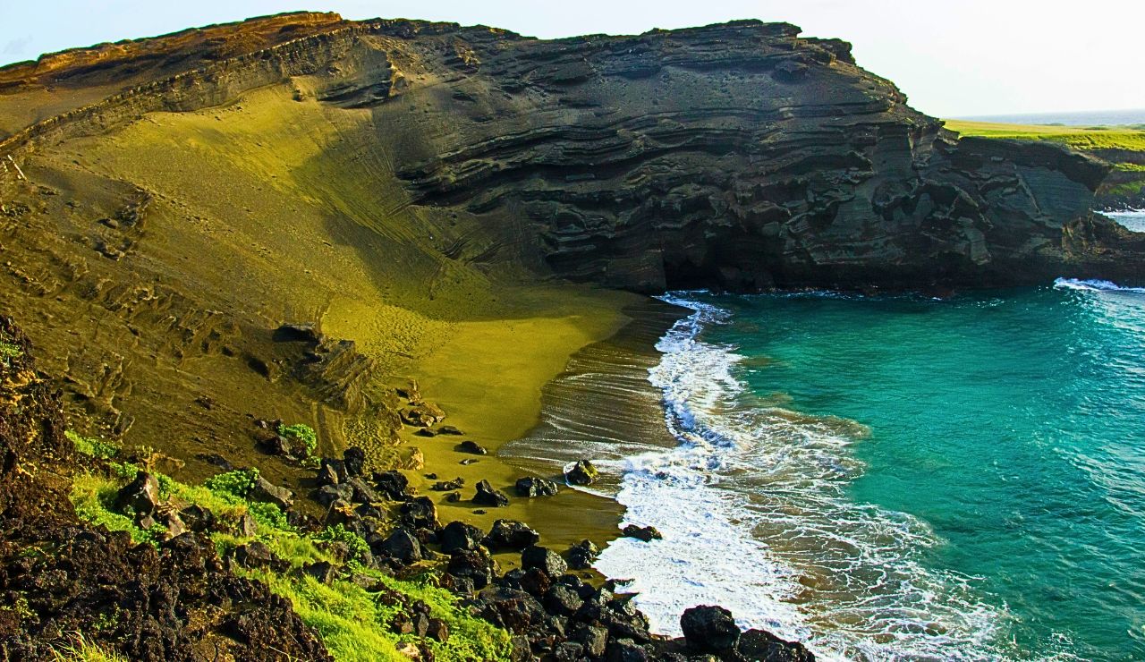 A high-angle view of Papakōlea’s olive-green sand curving around a turquoise bay, with a hiker in the distance.
