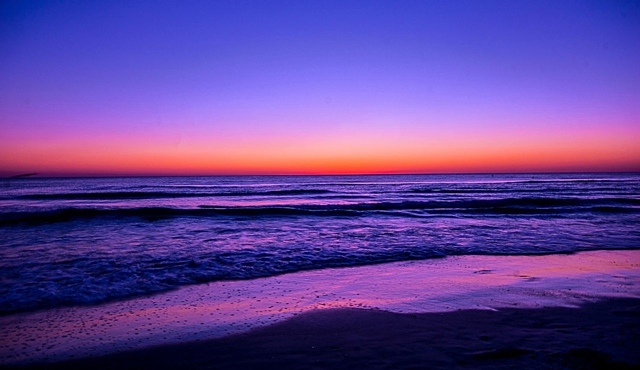 Purple-hued sand with a natural rock arch as waves crash through it at sunset.