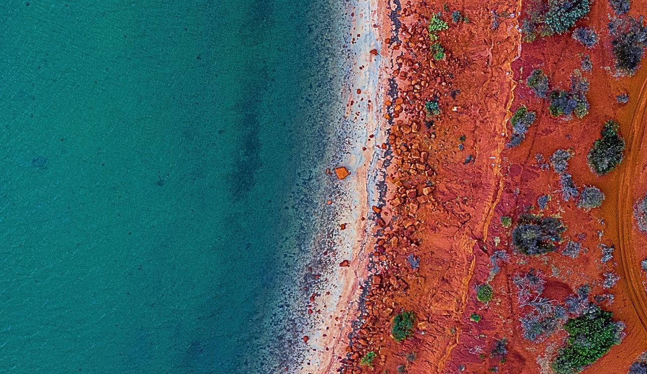 Crimson cliffs towering above a narrow red-sand shoreline and cobalt sea.