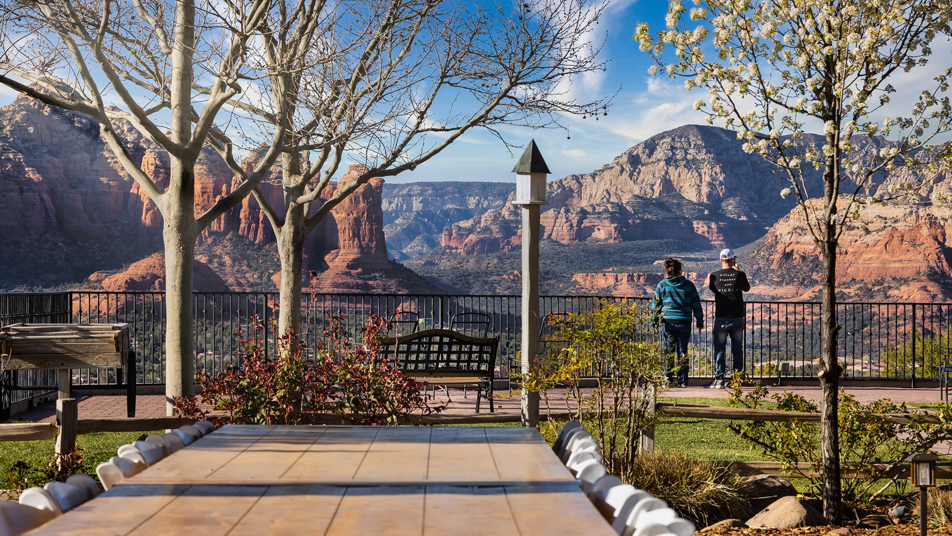 Two people stand by a railing overlooking red rock formations and mountains, with a table, chairs, and trees in the foreground.