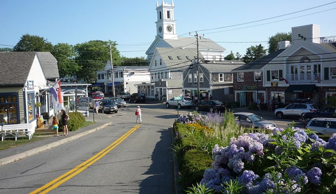 A charming street view of Chatham, Massachusetts, featuring a white clock tower, flower-filled sidewalks, and local shops under clear blue skies.