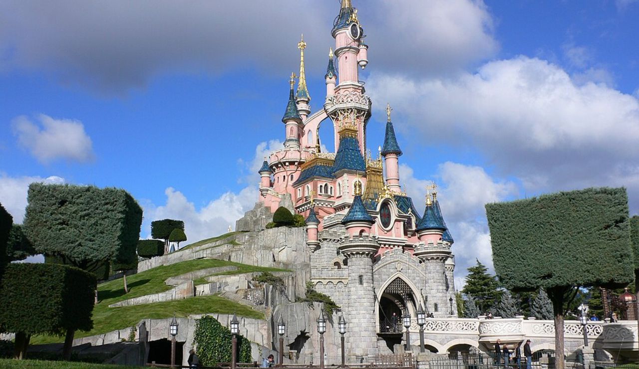 Fairy tale-style pink and blue castle with trimmed trees and stone pathway under a blue sky.