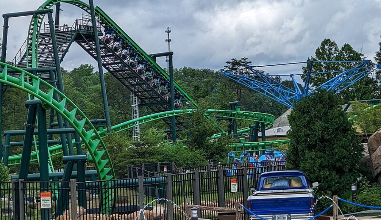 Green steel roller coaster with a steep climb and twists, set in a wooded amusement park with a blue vintage truck in the foreground.