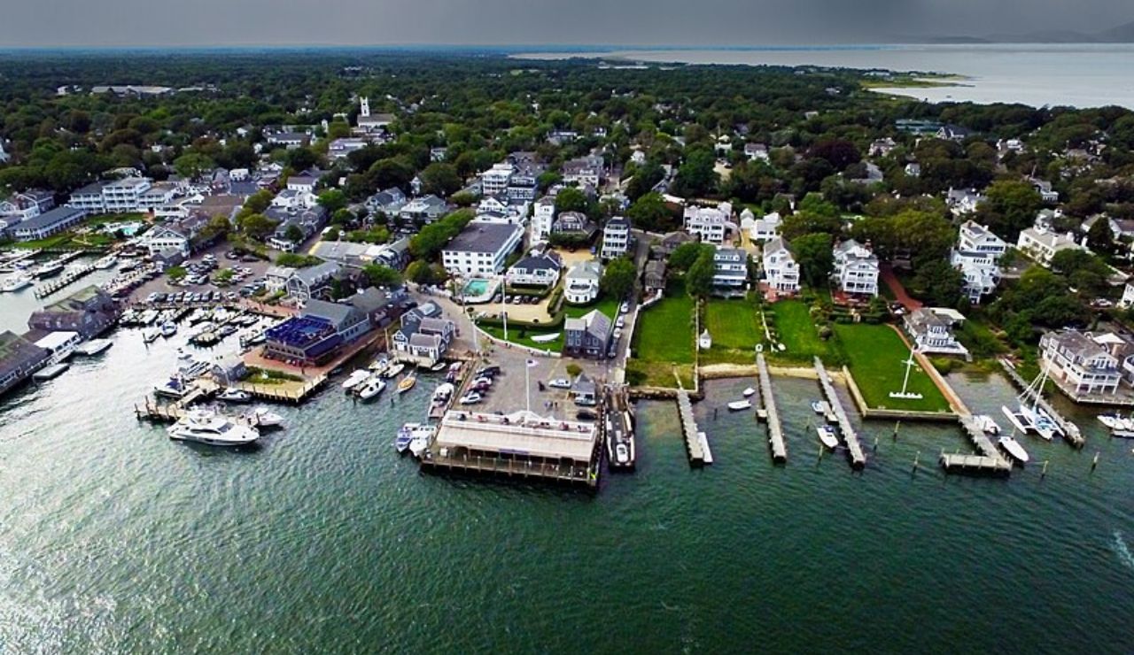 Aerial view of Edgartown, Massachusetts, showing a scenic harbor lined with boats, historic white homes, tree-filled neighborhoods, and a dramatic cloudy sky over the ocean horizon.