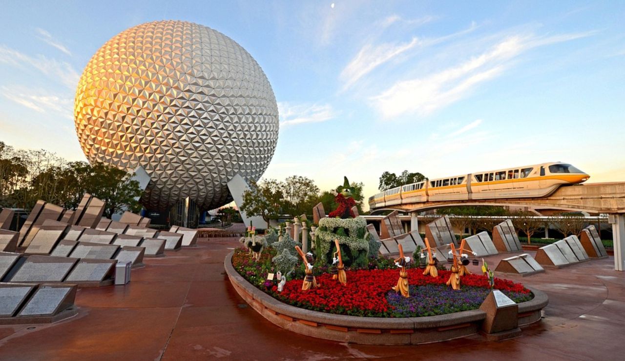 Geodesic sphere with a monorail passing behind flower sculptures and modern stone structures at a theme park entrance