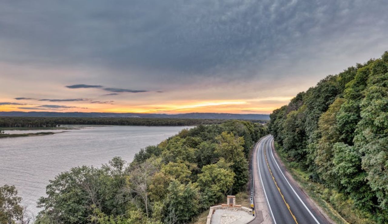 Winding road running alongside a river at sunset, bordered by dense green forest under a dramatic cloudy sky.