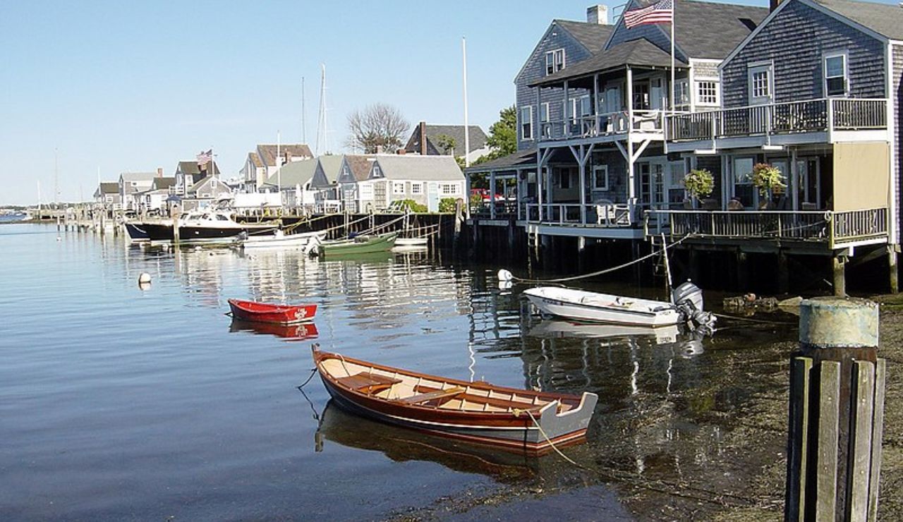 A peaceful harbor scene in Nantucket, Massachusetts, with wooden boats floating near charming gray-shingled seaside cottages and American flags fluttering in the breeze.