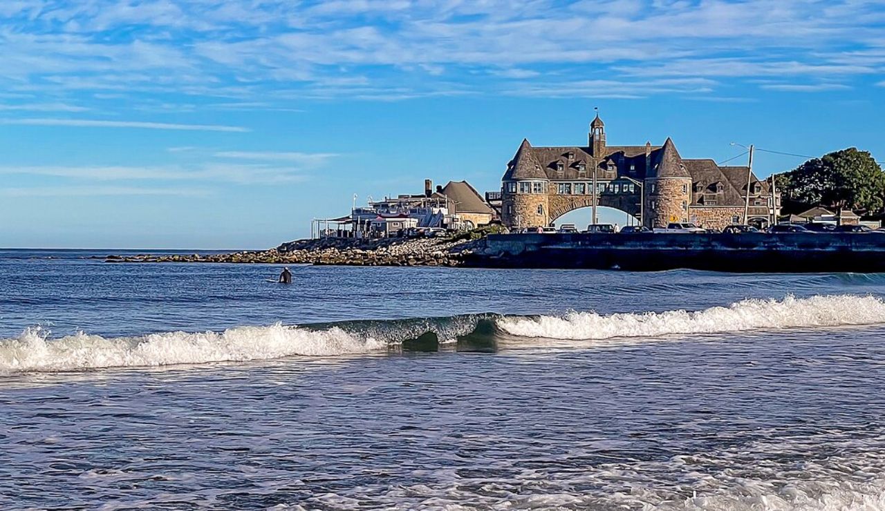 A stone towered building with an archway, known as The Towers in Narragansett, Rhode Island, overlooks gentle ocean waves under a bright blue sky.