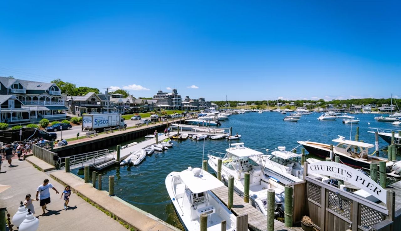 A sunny view of Church’s Pier in Oak Bluffs, Massachusetts, showing docked boats, waterfront homes, and people strolling along the marina under a bright blue sky.