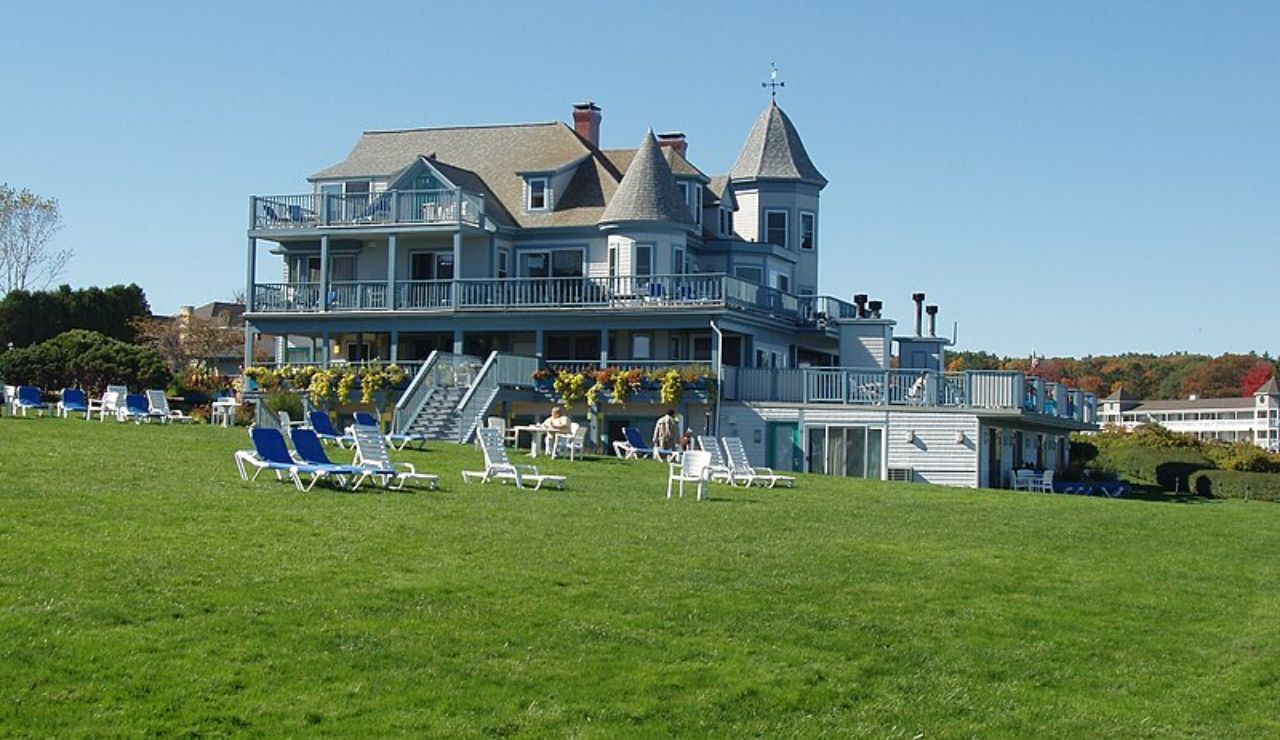 Large Victorian-style inn with wraparound porches and lawn chairs on a green grassy hill under a clear blue sky.