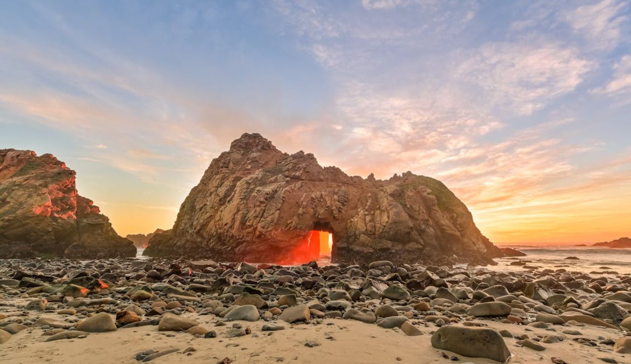 Sunset at Pfeiffer Beach in Big Sur, California, with sunlight streaming through a natural rock arch and illuminating the rocky shoreline.