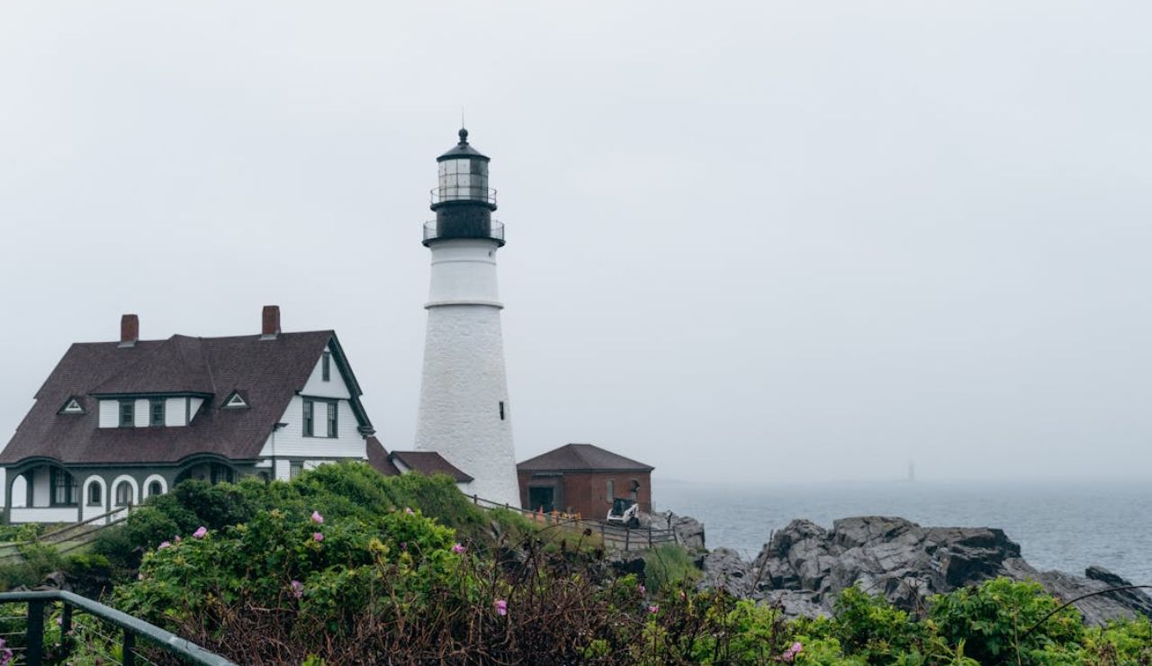 Portland Head Light in Maine on a foggy day, with a historic house beside it and waves crashing on rocky cliffs.